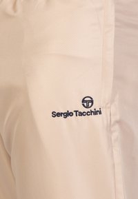 Camisa deportiva de un tono beige claro con un patrón texturizado, que presenta un logo en azul marino de "Sergio Tacchini" en el frente. Tejido suave, diseño casual.