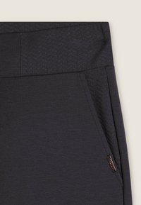 Pantalones deportivos gris oscuro con cintura texturizada, bolsillo lateral y un detalle metálico de la marca. Tela suave con ligero brillo.