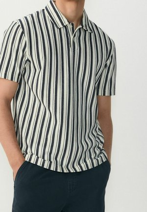 Homme portant un polo à manches courtes à rayures verticales noires et blanches, et un pantalon bleu foncé, les mains dans les poches, sur fond uni.