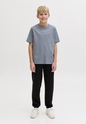 Ragazzo biondo in piedi e sorridente, indossa una maglietta grigia a maniche corte, pantaloni neri e sneakers bianche su sfondo bianco.