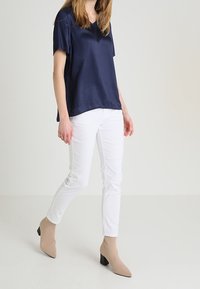 Kvinna som går, iförd en lös marinblå kortärmad topp, vita åtsittande byxor och beige ankelstövlar med klack.