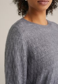 Grijze gebreide trui met een gestructureerd ruitpatroon en een ronde hals. Het materiaal lijkt zacht en lichtgewicht.