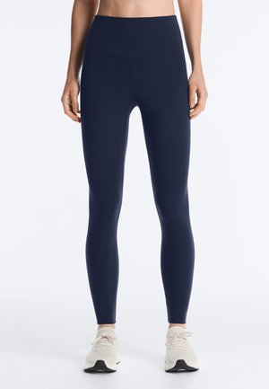 Žena oblečená v tmavomodrých legínach s vysokým pásom a bielych športových topánkach stojaca na jednoduchom bielom pozadí.