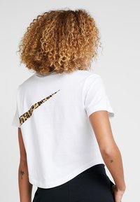 Bílý zkrácený tričko vyrobené z bavlny, s leopardím potiskem loga Nike Swoosh na zadní straně. Krátké rukávy a volný střih.