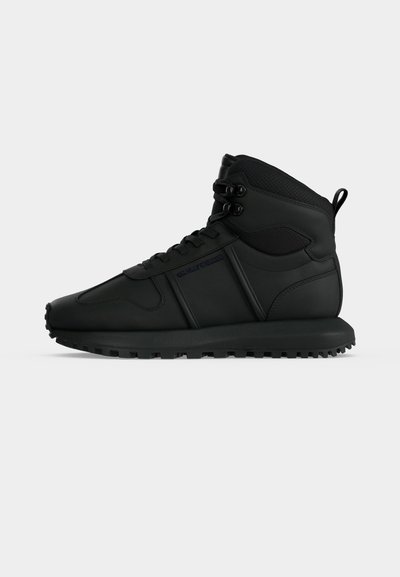 Μαύρο sneaker ψηλού τύπου με παχιά σόλα, δέσιμο μπροστά και ταινία έλξης στη φτέρνα, εμφανίζεται από το πλάι σε απλό λευκό φόντο.