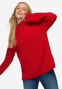 Rode gebreide trui met een geribbelde textuur, oversized pasvorm en zijkanten. Gepaard met blauwe spijkerbroeken.