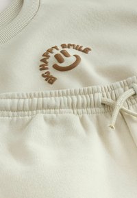 Beżowa bluza z haftowanym brązowym logo "BIG HAPPY SMILE". Posiada ściągacz przy okrągłym dekolcie oraz elastyczny pas z sznurkiem. Miękka w dotyku.