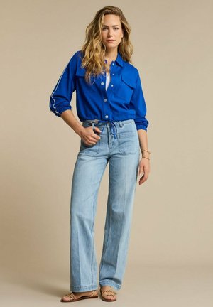 Vrouw staand, gekleed in een felblauw jasje met knopen, lichtblauwe wijde jeans en sandalen met luipaardprint tegen een beige achtergrond.