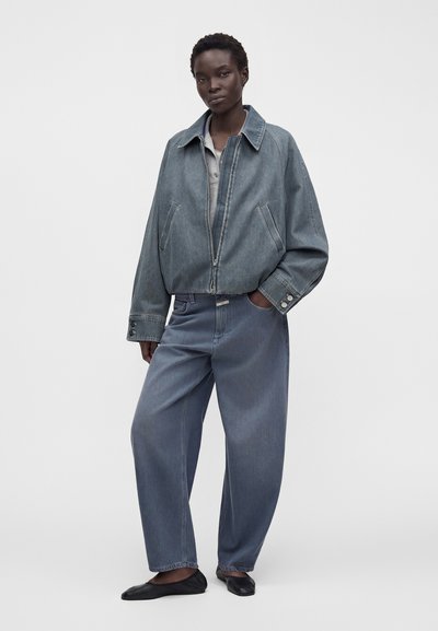 Femme portant une veste en jean bleue oversize et un jean à jambes larges avec des ballerines noires, debout avec une main dans la poche sur fond blanc.