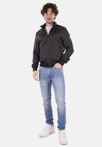 Giacca bomber in nylon nera con colletto alto, chiusura a zip, tasche laterali e orlo a costine. Indossata con jeans azzurri e sneakers bianche.