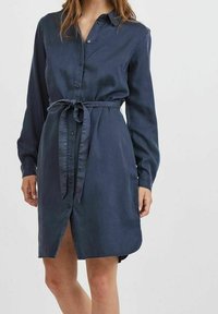 Denimblå skjolklänning med långa ärmar, knäppning fram, egen knytbelte i midjan och sidofickor. Slät textur och knä-lång klipp.