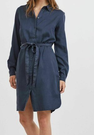 Abito camicia in denim blu con maniche lunghe, abbottonatura frontale, cintura da annodare in vita e tasche laterali. Tessuto liscio e lunghezza al ginocchio.