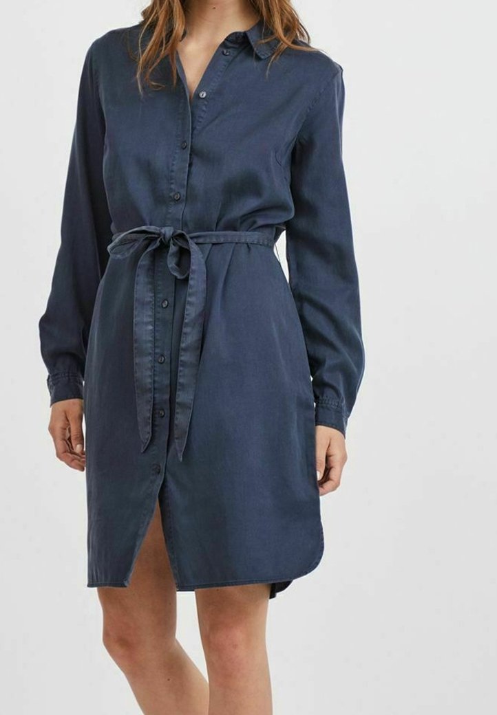 Denimblå skjolklänning med långa ärmar, knäppning fram, egen knytbelte i midjan och sidofickor. Slät textur och knä-lång klipp.