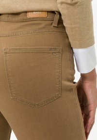 Pantalones de algodón marrones con un bolsillo trasero con un contorno cosido y sutiles adornos, combinados con una blusa beige y una manga blanca.