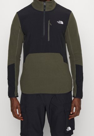 Mężczyzna ubrany w oliwkowo-zielony i czarny polar z suwakiem na kieszeni na piersi oraz czarne spodnie typu cargo, oba z logo The North Face.