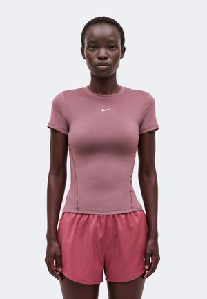 Žena s krátkymi vlasmi nosiaca priliehavé fialové tričko Nike a voľné ružové šortky stojaca pred jednoduchým svetlým pozadím.