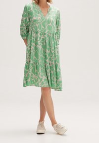 Kobieta w zielonej i białej sukience w liście do kolan, z rękawami trzy czwarte i białymi trampkami, stojąca z nogami skrzyżowanymi.