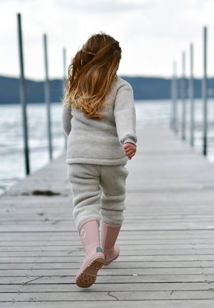 Pulover gri din fleece și pantaloni asortați, cu cizme roz din cauciuc. Cizmele au un logo și tălpi texturate, văzute din spate pe un ponton de lemn.