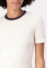 Kobieta ubrana w biały, prążkowany t-shirt z krótkim rękawem, z granatowo-czerwono-białym kołnierzykiem na tle czystej białej ściany.