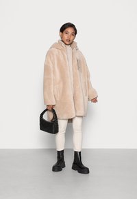 Manteau oversize en fausse fourrure beige avec une capuche, des poches latérales et un détail de panneau beige. Associé à des leggings blancs et des bottes noires à la cheville.