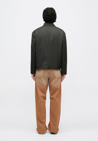 Zwarte, op maat gemaakte jas met een gladde textuur, gecombineerd met bruine, vervaagde brede pijpen spijkerbroek met achterzakken. Achteraanzicht.