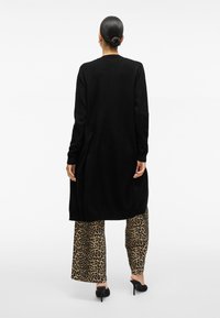 Svart lång cardigan med ribbad kant och muddar, kombinerad med vida byxor i leopardmönster och svarta öppna klackskor. Bakifrån.