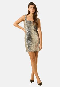 Robe mini en sequins argentés avec décolleté carré et bretelles épaisses. Texture lisse avec une silhouette ajustée. Associée à des talons hauts noirs.