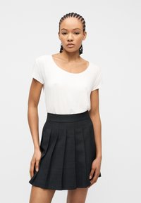 Γυναίκα με πλεξούδες, φοράει λευκό τοπ με κοντά μανίκια και μαύρη φούστα με πιέτες και ψηλή μέση, στέκεται μπροστά σε απλό φωτεινό φόντο.