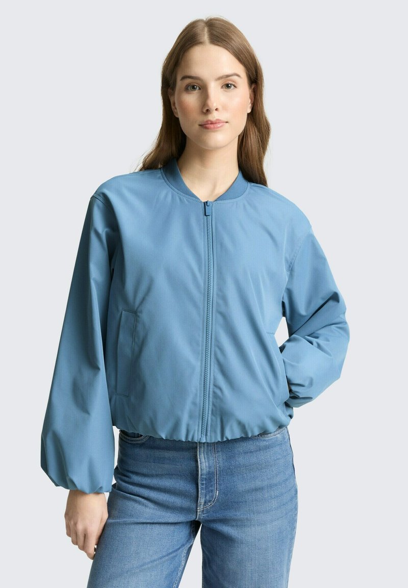Frau mit glattem hellbraunem Haar, die eine hellblaue Bomberjacke mit Reißverschluss und blaue Jeans trägt, steht mit einer Hand in der Hüfte.