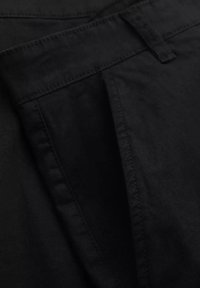 Pantalon noir en coton lisse, avec un devant plissé et des coutures contrastantes. La taille est dotée d'une fermeture à bouton.