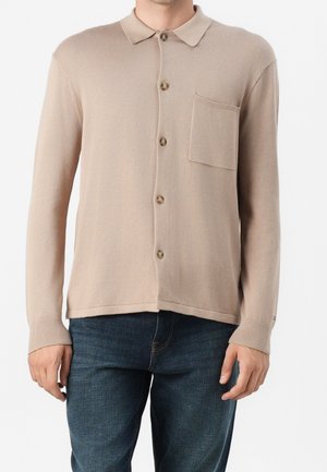 Beige long-sleeve buttoned shirt with collar and chest pocket worn with dark blue jeans by a standing person.