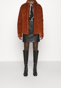 Braune Cord-Jacke mit gepolstertem Muster, zwei Brusttaschen, über einem schwarzen Shirt und einem Lederrock getragen. Schwarze knielange Stiefel mit Absatz.