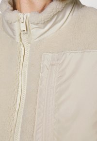 Krämsfärgad jacka med ett mjukt, texturerat, fleece-liknande material och släta, glänsande paneler samt en hög krage. Dragkedja fram.