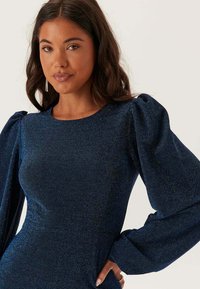 Mörkblå glittrig klänning med långa puffärmar, rund halslinje och en figurnära siluett, som visar en slät, texturerad tyg.