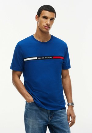 Ung mand iført en blå Tommy Hilfiger T-shirt med logo og røde, hvide, sorte striber, blå jeans og et sølv armbåndsur.