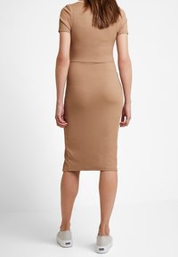 Kobieta stojąca tyłem, ubrana w dopasowaną, prążkowaną, beżową sukienkę midi z krótkim rękawem oraz białe sportowe trampki na białym tle.