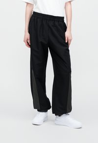 Schwarze Sporthose mit weit geschnittenem Bein, elastischem Bund und kontrastierenden dunkelgrünen Seitenpanelen. Kombiniert mit weißen Turnschuhen.