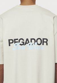 Krēmkrāsas t-krekls ar īsām piedurknēm. Aizmugurē ir uzraksts „PEGADOR” treknrakstā melnā krāsā un švītīgā zilos burtiņos „Next Wave”. Gluda tekstūra.