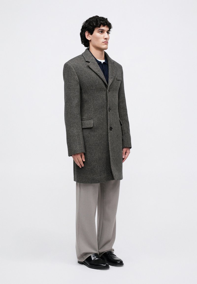 Homme debout dans un manteau ajusté marron, un pantalon large gris, des chaussures noires et un pull sombre sur fond clair. 