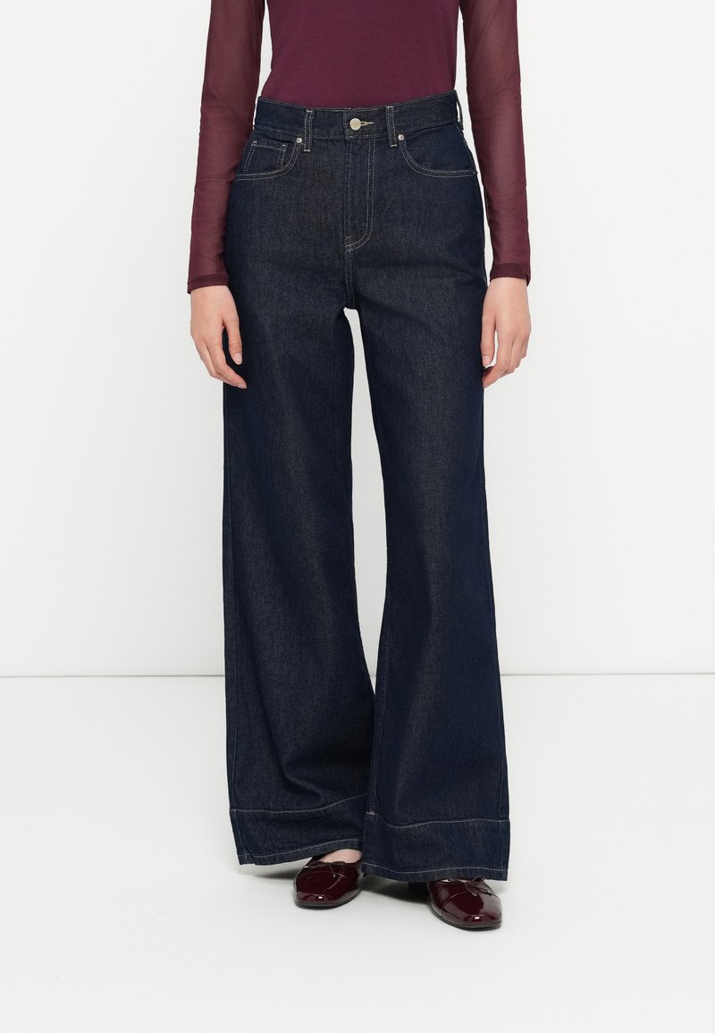 Høyt liv, vide ben i mørk denim med to frontlommer, diskrete sømdetaljer og knappelukking. Kombinert med en bordeaux topp.