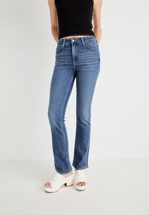 Jeans de mezclilla azul con un corte ajustado y piernas ligeramente acampanadas, con cinco bolsillos y cierre de botón. Combinados con tacones de plataforma blancos.