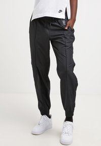 Black track pants made of smooth fabric, featuring side pockets, elastic waistband, and snap buttons along the side. Paired with white sneakers.