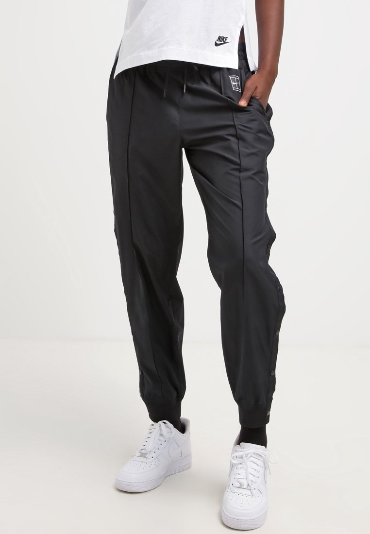 Black track pants made of smooth fabric, featuring side pockets, elastic waistband, and snap buttons along the side. Paired with white sneakers.