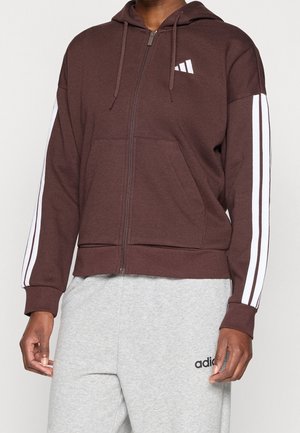 Hombre que lleva una sudadera marrón con cremallera y rayas blancas en las mangas y pantalones deportivos grises con un logo negro en la pierna izquierda.