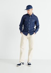 Camisa de mezclilla de un azul oscuro con dos bolsillos en el pecho, pantalones color crema y zapatillas marinas. La gorra coincide con el color de la camisa. Diseño simple.