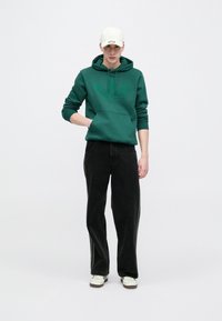 Πράσινο φούτερ με κουκούλα και μπροστινή τσέπη, συνδυασμένο με μαύρο παντελόνι φαρδιάς γραμμής. Λευκό καπέλο και λευκά αθλητικά παπούτσια που ταιριάζουν ολοκληρώνουν το σύνολο.