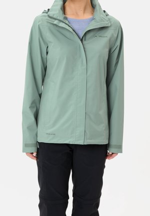 Personne portant une veste imperméable vert clair avec une capuche et un pantalon noir sur un fond uni clair.