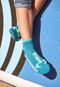 Die Beine eines Kindes tragen blaue Anti-Rutsch-Socken mit dem Text "BEANSM" und sitzen auf einer blauen und schwarzen strukturierten Matte im Freien.