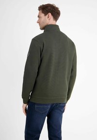 Dunkelgrüner Reißverschluss-Pullover mit hohem Kragen, strukturierter Stoff und gerippten Bündchen, kombiniert mit blauen Jeans. Keine sichtbaren Logos oder Muster.
