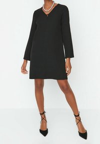 Kvinna som bär en svart långärmad klänning med V-hals, silverpärlhalsband och svarta spetsiga klackskor med ankelremmar, stående mot en vit bakgrund.